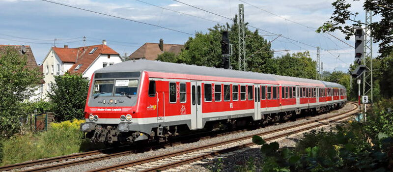 Roter ÖPNV fährt auf Gleisen durch grüne Landschaft mit Bäumen, Häsern und Stromleitungen.