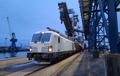 Eine weiße WFL-Lok-BR 248 Vectron Dual mit angehängten Güterwagen steht zum beladen an einer Hafenanlage. Zu sehen sind mehrere Beladungskräne und eine Anlage mit Förderband für Schüttgut. Es ist Abenddämmerung.