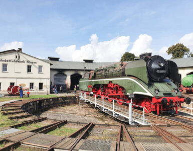 Großansicht einer schwarz-grünen WFL Dampflok mit rotem Fahrwerk Modell 020201-0  auf dem Drehplatz im Bahnbetriebswerk Nossen. Im Hintergrund sind Besucher zu sehen, sowie das Werkstattgebäude.