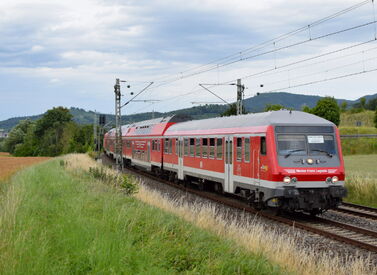 Roter ÖPNV Doppelstockzug fährt auf Gleisen durch grüne Landschaft mit Bäumen, Wiese und Stromleitungen.