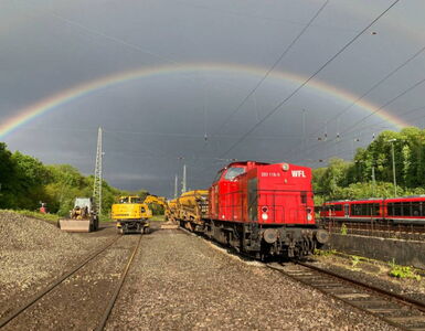Stimmungvolles Bild mit Regenbogen über einer Gleisstrecke. Bagger erneuern das Gleisbett mit Schottersteinen. Eine rote WFL Diesellokomotive mit angehängten Güterwagons fährt vorbei..