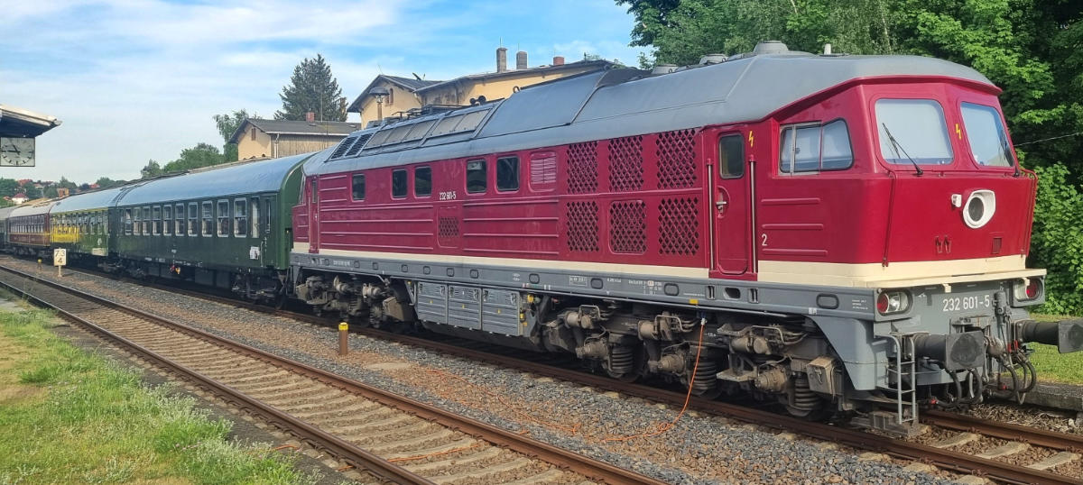 rote Diesellok 232 mit angehängten historische Reisezugwagen bei Vorbeifahrt an einem ländlichen Bahnhof