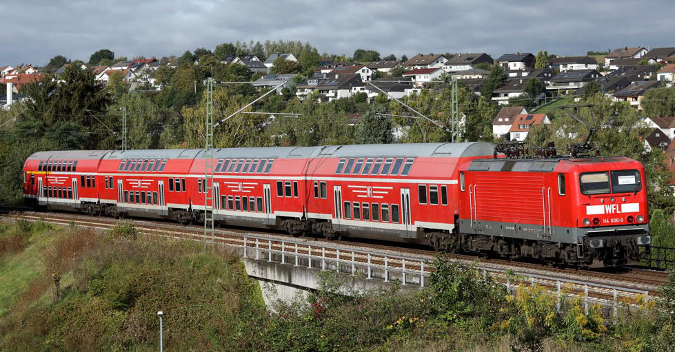 Roter WFL ÖPNV Doppelstockzug fährt durch Landschaft über eine kleine Brücke. Im Hintergrund sieht man eine Kleinstadt.